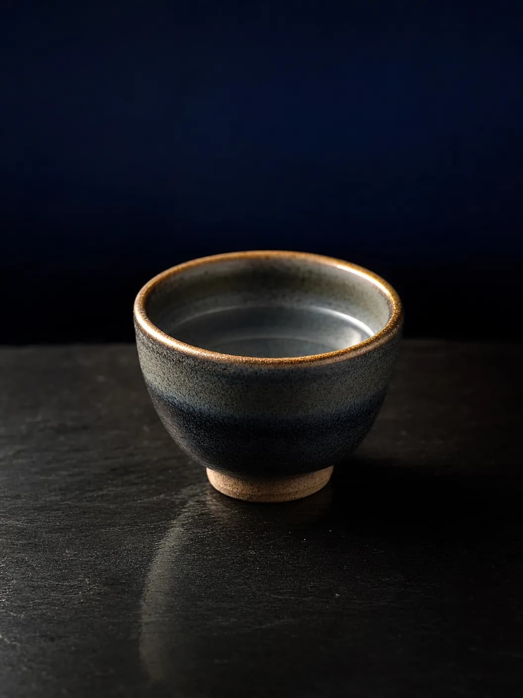 Close-up of a ceramic ochoko sake cup with warm gold rim light against deep navy